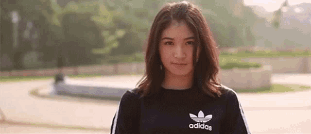 a woman wearing a black adidas shirt is standing in a park and looking at the camera .