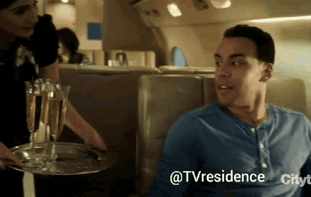 a man sitting on a plane with a tray of champagne glasses and the words @tvresidence city on the bottom