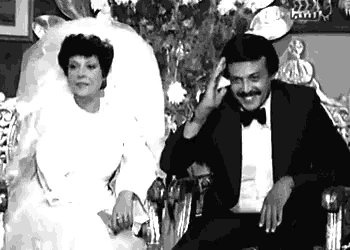a black and white photo of a bride and groom sitting next to each other on a chair .
