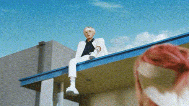 a man in a white suit is sitting on the roof of a building next to a woman with pink hair .