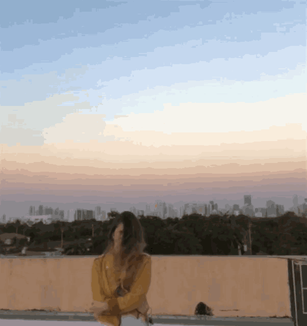 a woman in a yellow sweater is standing on a rooftop with a city skyline in the background