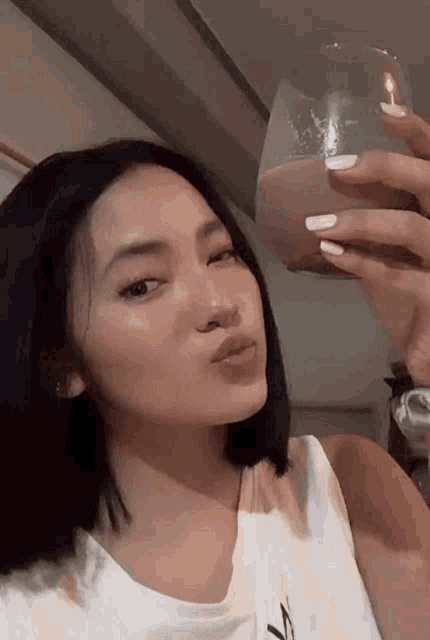 a woman in a white tank top is holding a glass of liquid in her hand .