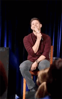 a man in a red shirt is sitting in front of a crowd holding a microphone and smiling