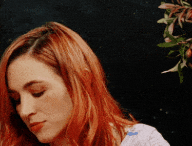 a woman with red hair looks down with a flower in the background