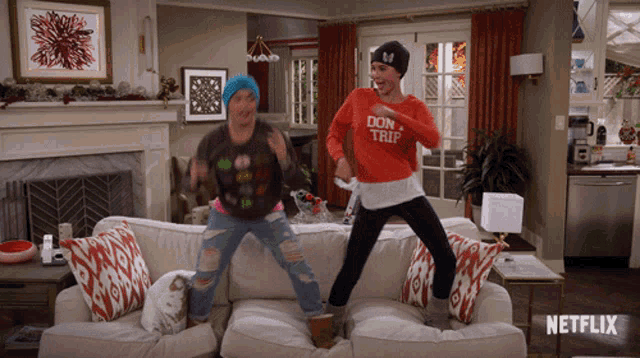 two women jumping on a couch with one wearing a sweater that says don 't trip