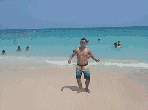a shirtless man stands on the beach giving a peace sign