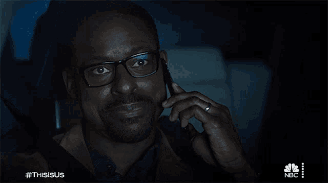a man wearing glasses is smiling while talking on a cell phone with a nbc logo in the background