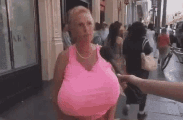 a woman with very large breasts in a pink tank top is being interviewed on a street .