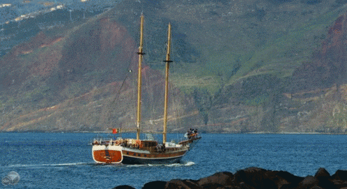 a sailboat is floating in the ocean with mountains in the background and a bbc logo in the corner