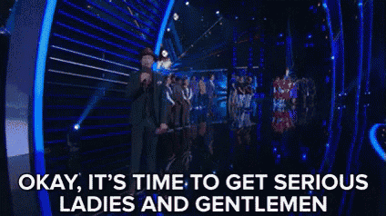 a man in a suit stands on a stage with a microphone and says okay it 's time