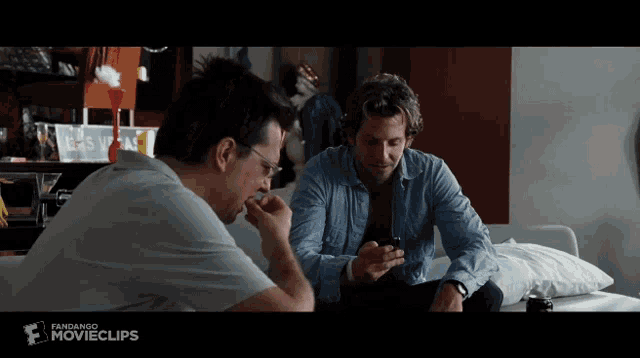 two men are sitting on a couch and one is looking at his phone