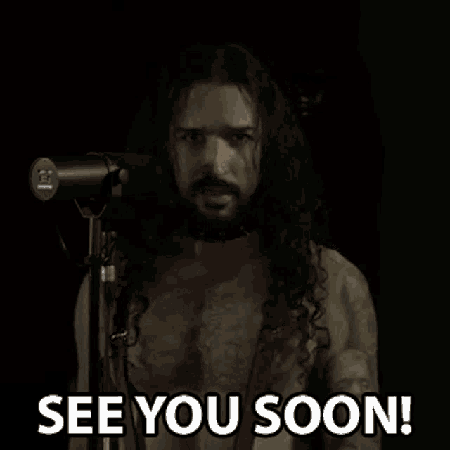 a man with long hair and a beard is standing in front of a microphone and says `` see you soon '' .