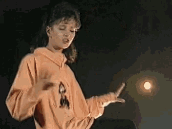 a woman in an orange sweatshirt is standing in front of a light and making a gesture with her hands .