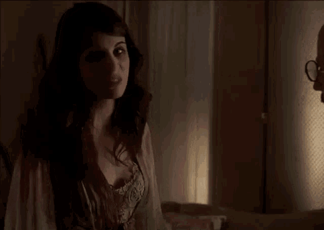a woman is standing in front of a window in a dark room talking to a man .