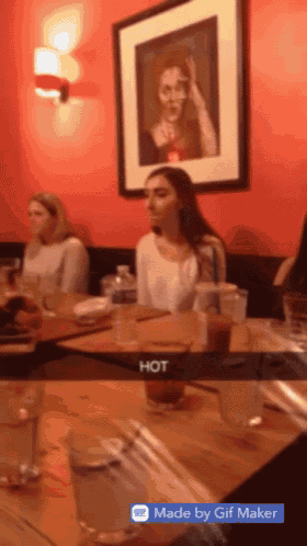 a snapchat of a woman sitting at a table with a picture on the wall that says hot