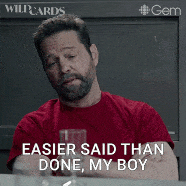 a man in a red shirt is sitting at a table with a glass of wine and says " easier said than done my boy "