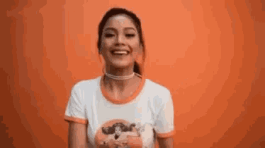a woman in a white t-shirt and choker is smiling in front of an orange wall .