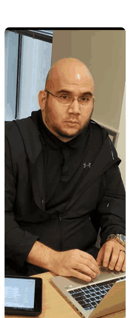 a man wearing glasses and a black under armour jacket is typing on a laptop