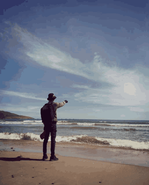 a man in a hat stands on a beach pointing at the ocean