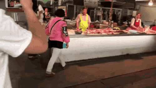 a woman in a pink jacket is walking in front of a butcher 's counter