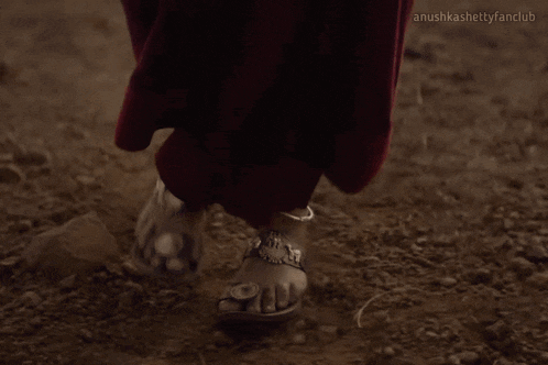 a woman in a red dress is walking on a dirt road with the words anushkashettyfanclub below her