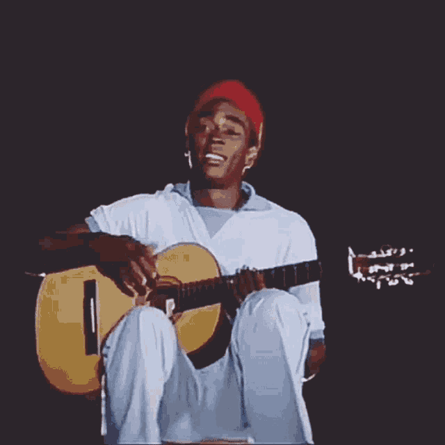 a man wearing a red hat is sitting down playing a guitar .