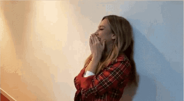 a woman in a plaid shirt is leaning against a wall and covering her mouth with her hands .