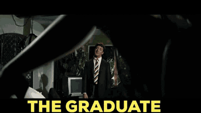 a man in a suit and tie is standing in a room with the words the graduate behind him