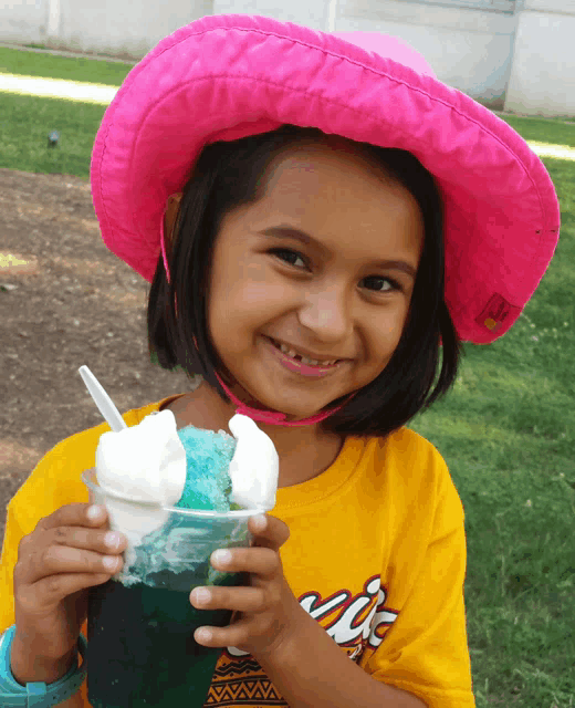 a young girl wearing a pink hat and a yellow shirt holds a cup of ice cream