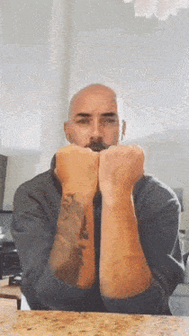 a bald man with a tattoo on his wrist is sitting at a table with his hands folded .