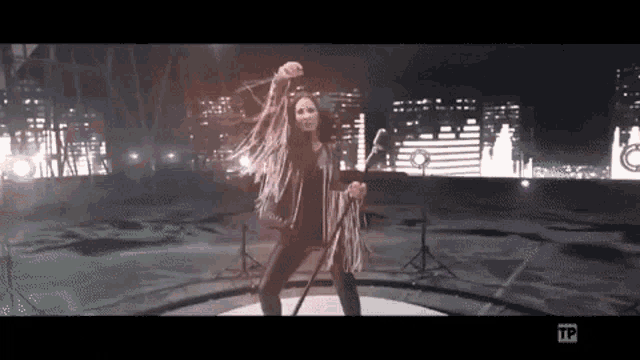 a woman in a fringed jacket is singing into a microphone in front of a city skyline