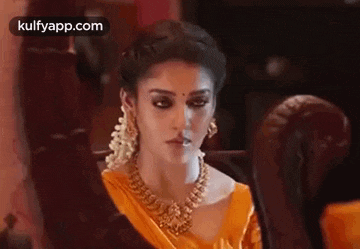 a woman in a yellow saree and gold jewelry is sitting on a couch with her hands in the air .