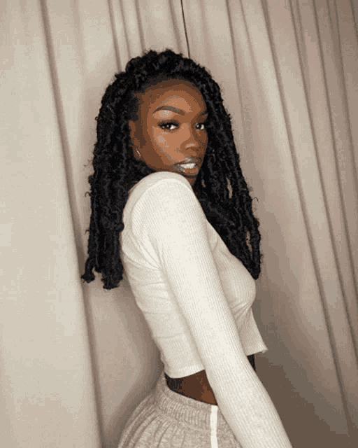 a woman with dreadlocks is wearing a white crop top and grey shorts