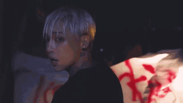 a man with white hair and a black shirt is standing in front of a wall with blood on it .