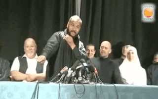 a man is standing at a table with a microphone in front of a crowd of people .
