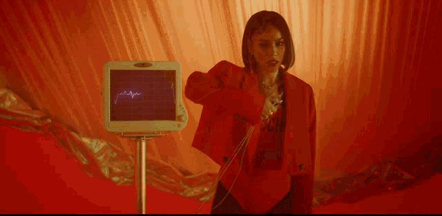 a woman in a red jacket is standing in front of a monitor with a heartbeat on it