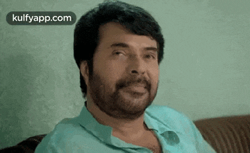 a man with a beard is sitting on a couch and looking at the camera .