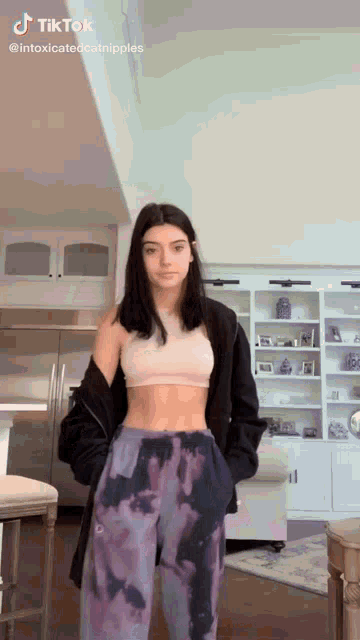 a woman is standing in a living room wearing a crop top and tie dye sweatpants .