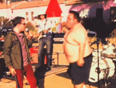 a man talking into a microphone while another man stands behind a drum set