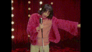 a woman in a pink fur coat is singing into a microphone on stage .
