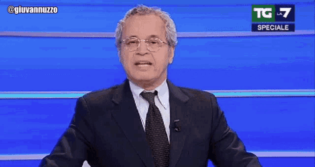 a man in a suit and tie is standing in front of a blue screen that says tg 7