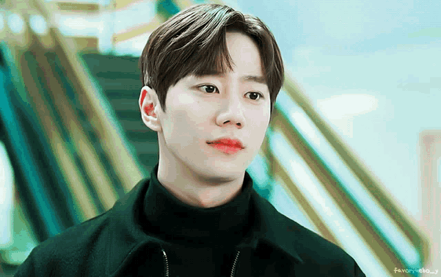 a young man wearing a black turtleneck looks at the camera in front of stairs