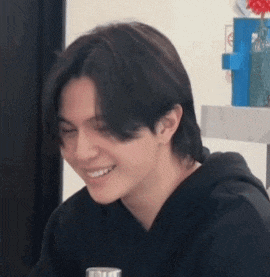a young man with long hair is smiling while sitting at a table with a glass of water .