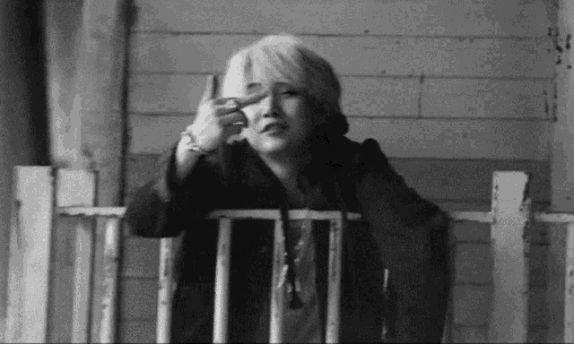 a black and white photo of a woman sitting on a porch giving the peace sign .