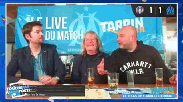 three men are sitting at a table in front of a live du match sign