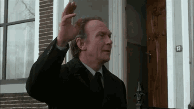 a man in a suit and tie is waving in front of a house with the number 13 on the door