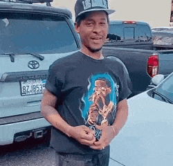 a man wearing a hat and a black t-shirt is standing in front of a car .