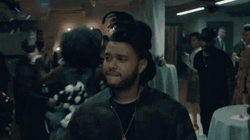 a man wearing a camouflage jacket and a necklace is standing in a room with a crowd of people .