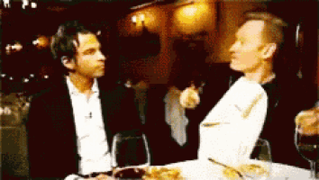 two men are sitting at a table with wine glasses and talking to each other