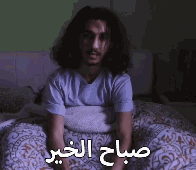 a man with long hair is sitting on a bed with the words " صباح الخير " written on the bottom